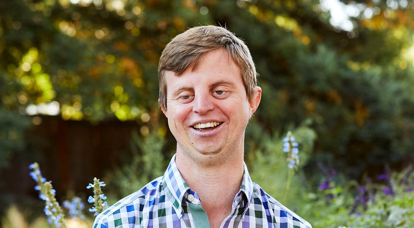 Dr. Hoby Webler is smiling at the camera and wearing a blue plaid shirt in front of a forest tree setting