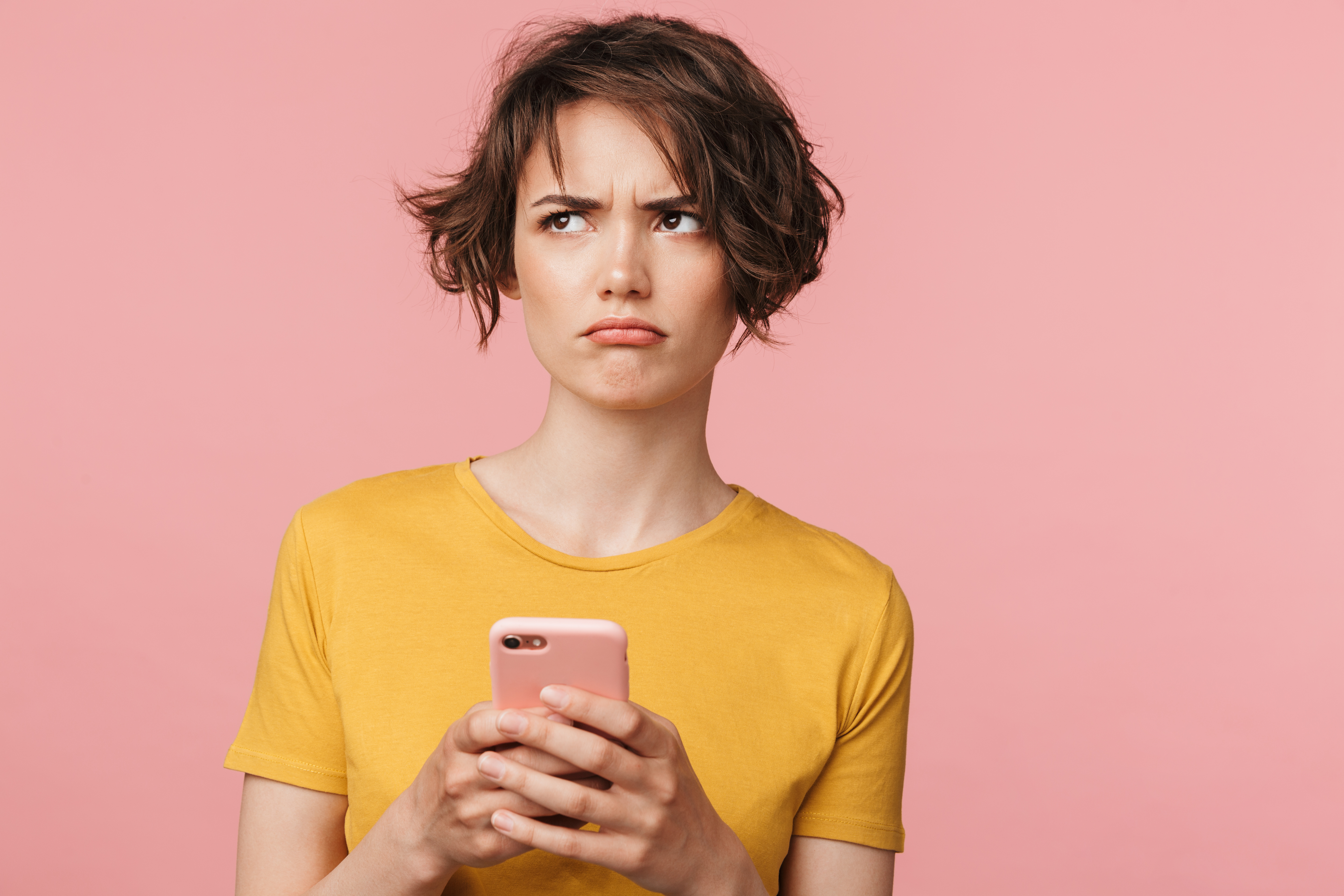 A brown-haired woman in a yellow shirt holding a mobile phone and she is looking to the side with a confused expression