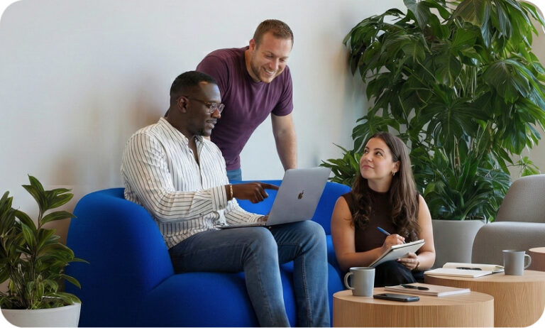 Three colleagues collaborating in a bright office lounge area. One person sits on a blue sofa with a laptop, a second person stands behind leaning in to look at the screen, and a third sits nearby taking notes in a notebook.