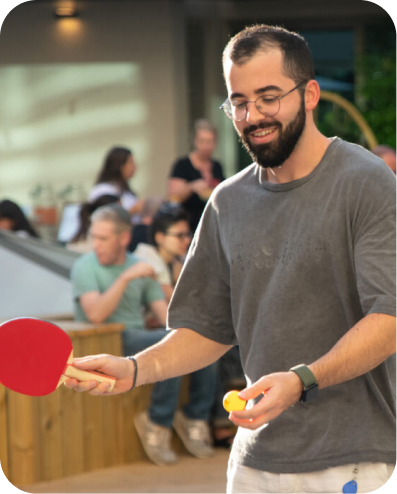 A presenter stands at the head of a conference room table, gesturing toward a large screen displaying a welcome slide. Eight colleagues sit around the table listening attentively.