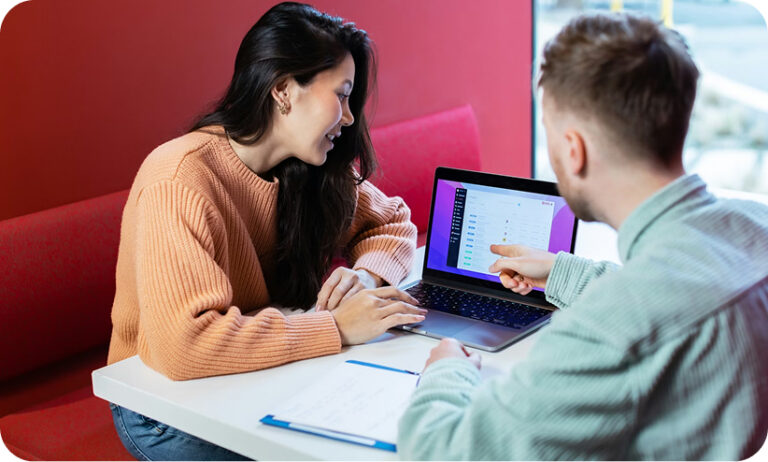 Two colleagues sitting at a table in a red booth, reviewing a dashboard on a laptop screen together. One person points at the screen while the other smiles and looks on.