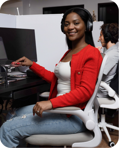A smiling woman wearing headphones and a red cardigan sits at a desk in an office, turned toward the camera with her hand on a computer mouse. A colleague wearing a headset is visible working in the background